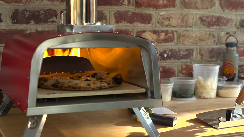 A pizza bakes in a small red outdoor oven on a countertop, with various ingredients in plastic containers beside it.