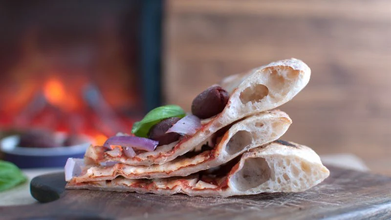 Three folded flatbread pizza slices with olives and red onions on a wooden surface, blurred fire in the background.