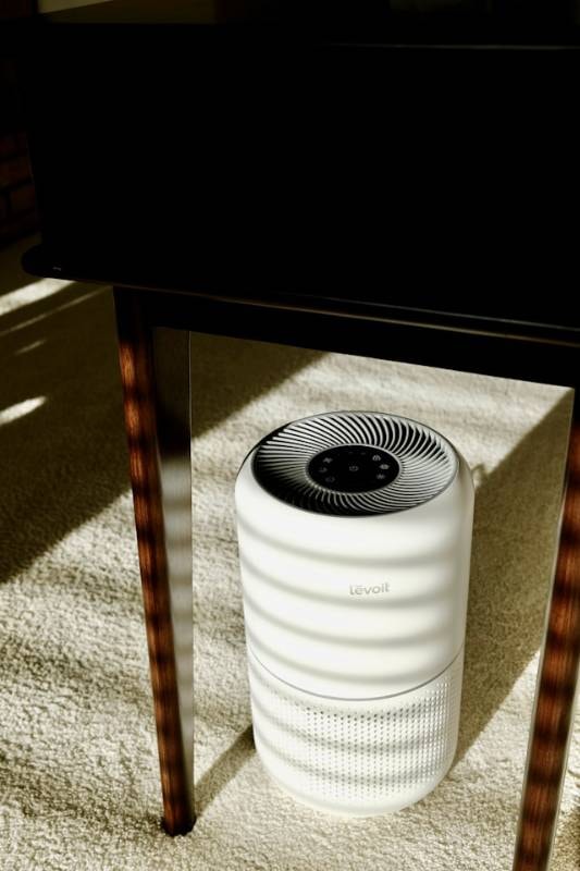 A white Levoit air purifier placed under a wooden table in a sunlit room, emphasizing the Health Benefits of Air Purifiers for Dogs