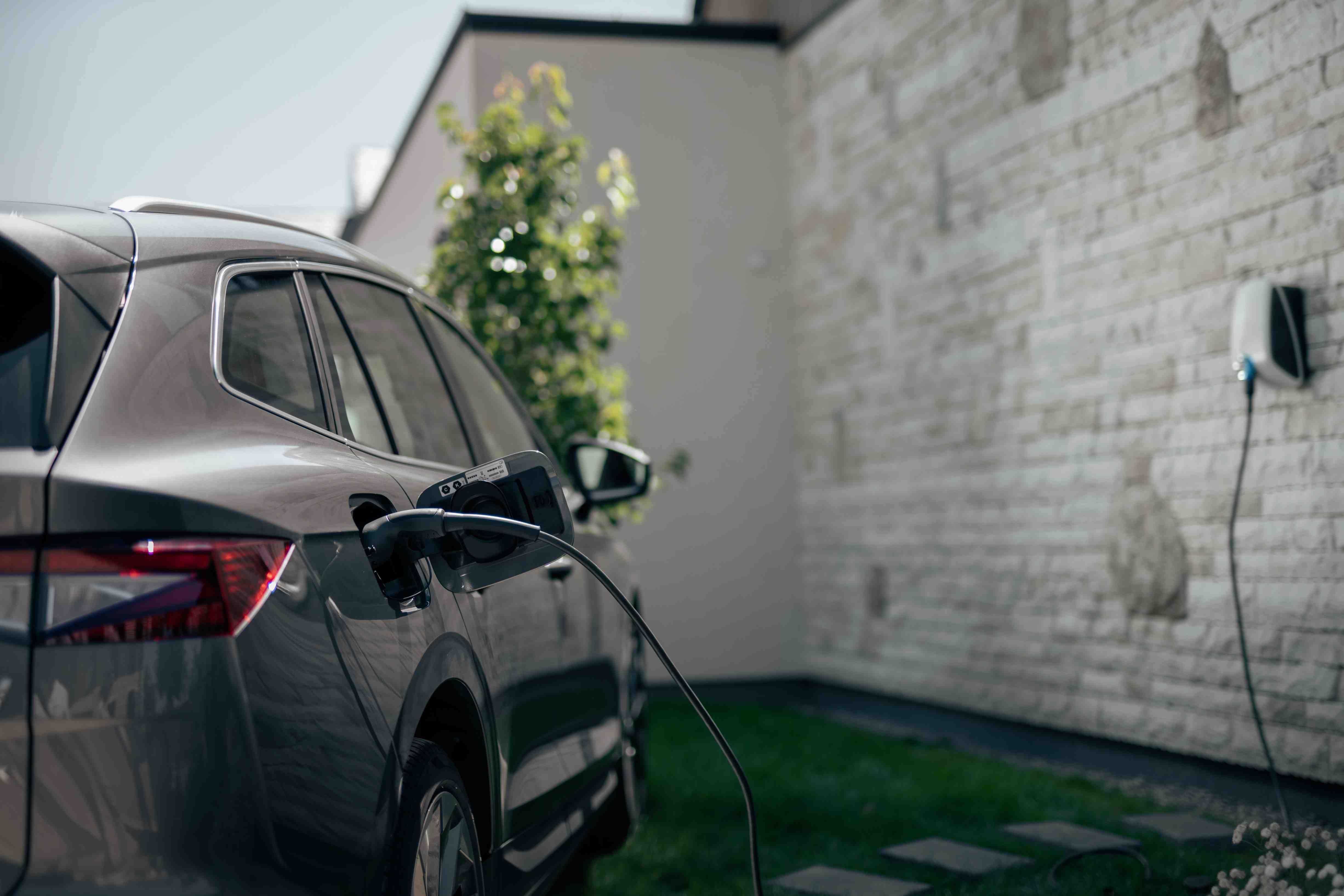 An electric SUV parked in the driveway and connected to a home charging station mounted on a brick wall.