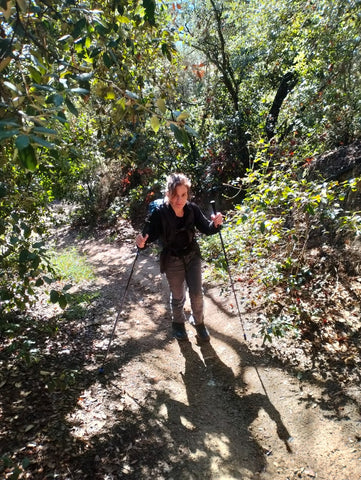Sarah, a white woman with brown hair wearing a dark long-sleeve shirt and jeans, is using her trekking poles on a hike through trees in Spain.