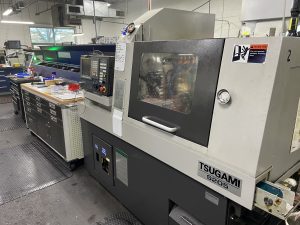 A Tsugami S205 Swiss Turn machine operating in a bustling manufacturing workshop filled with tool cabinets and various machining equipment.