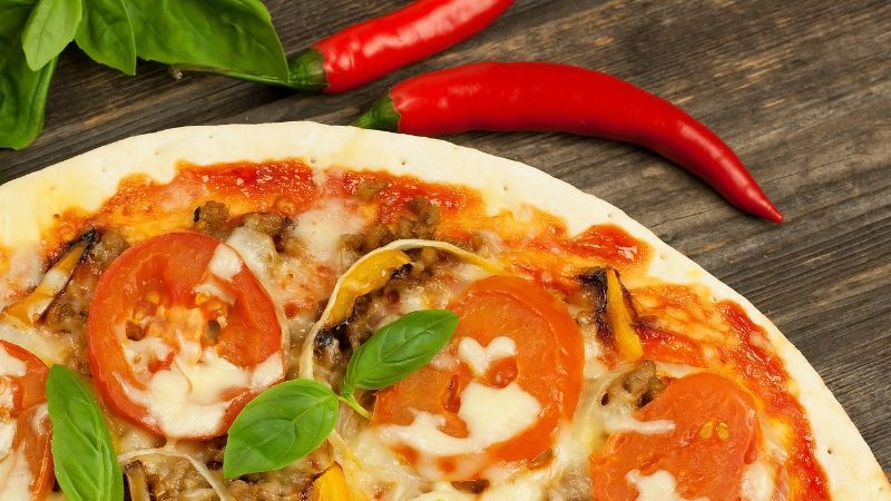 Close-up of a pizza with tomatoes, cheese, basil; two chili peppers and basil leaves on a wooden surface beside it.