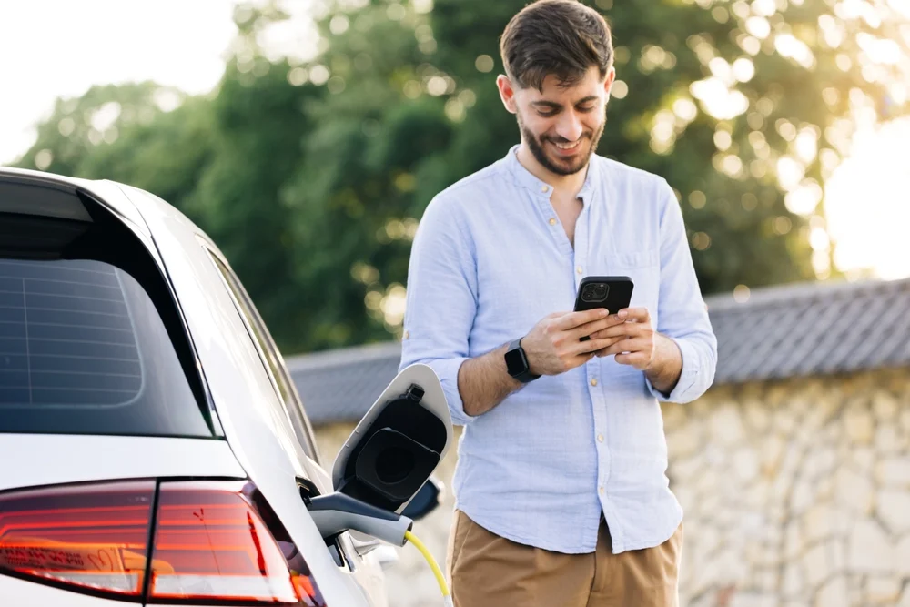A casual photo of a man checking his phone while his electric vehicle is charging.
