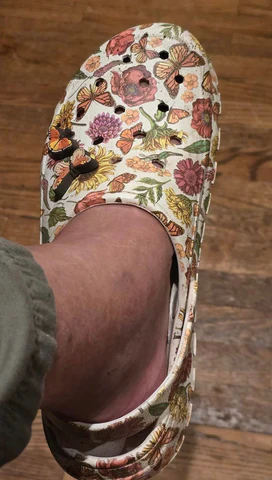 A person's foot wearing a croc-style clog that is white with a bold and colorful print of flowers and butterflies on them.
