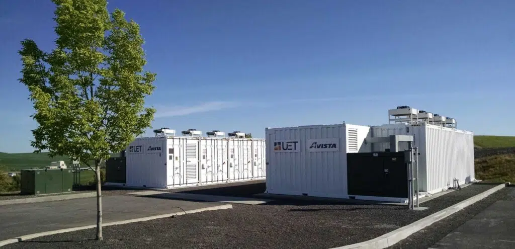 Two-part battery energy storage system surrounded by asphalt, grass, and a tree in the foreground