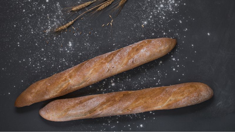 Two baguettes on a flour-dusted dark surface, with wheat stalks in the top left corner.