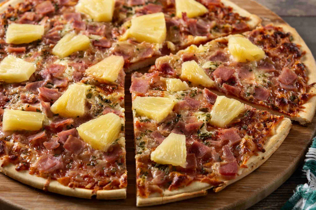 A partially sliced Hawaiian pizza with pineapple and ham on a wooden board.