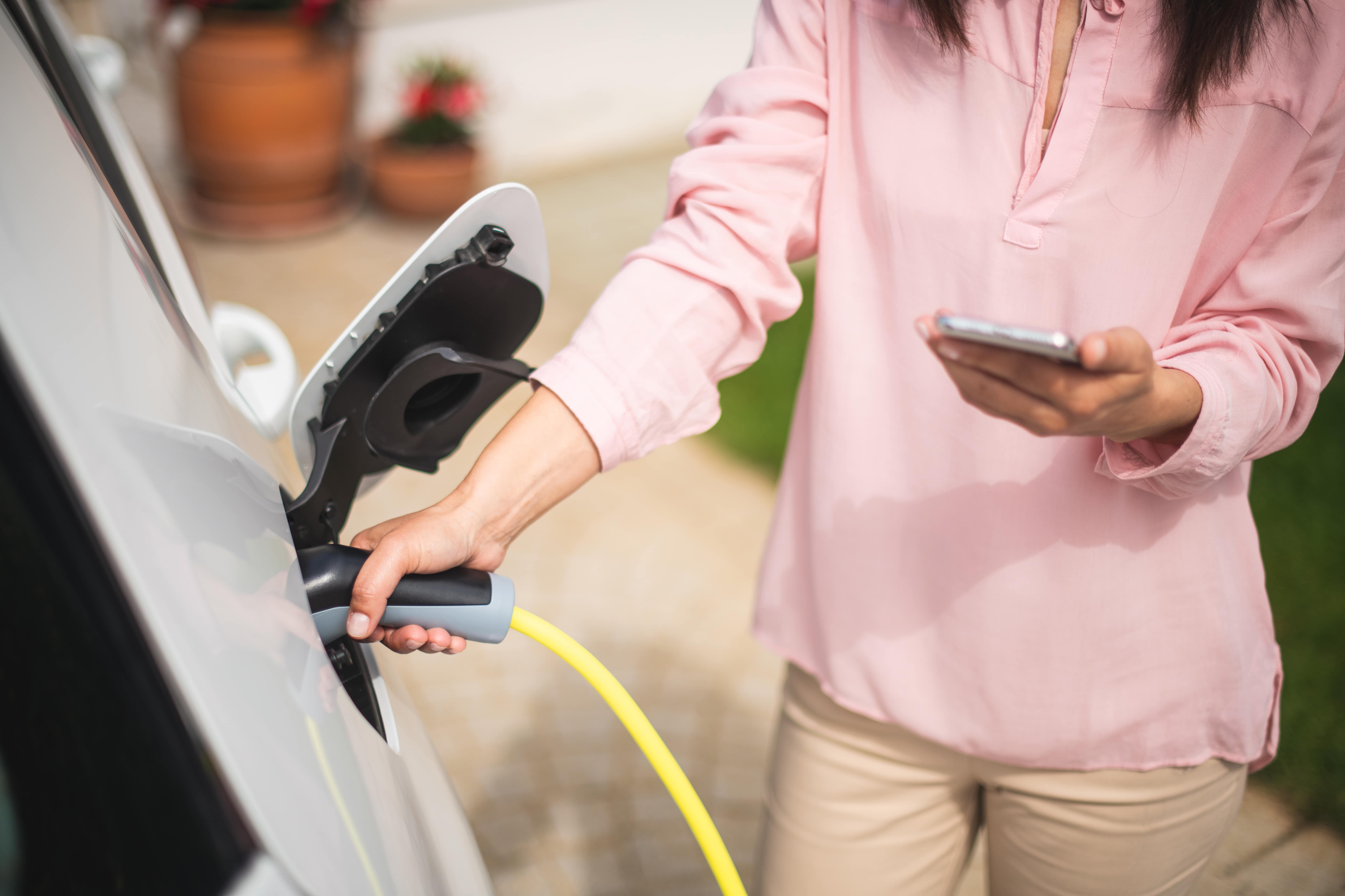 A close-up photo of a woman plugging in her electric vehicle while holding her smartphone.