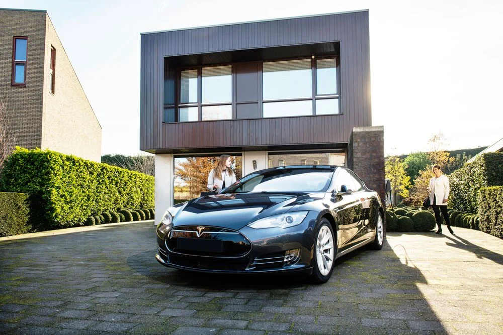 A black electric car is parked in front of a modern house on a sunny day.