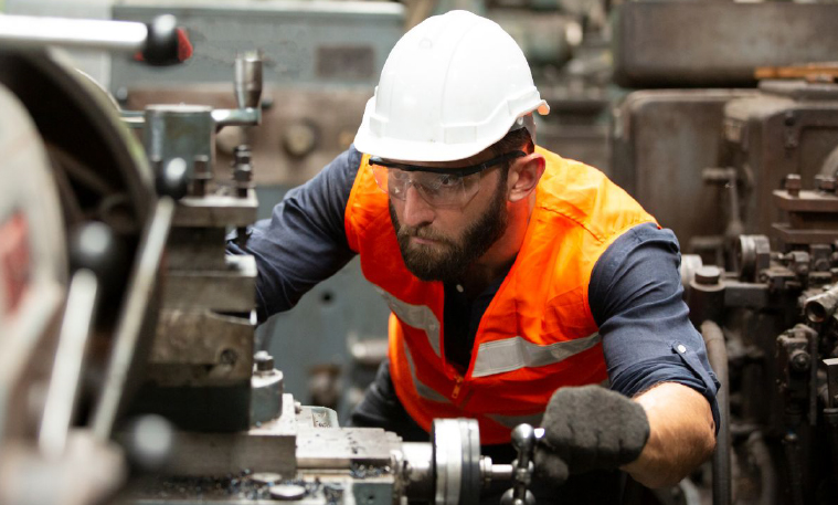 Industrial worker wearing a white hard hat, safety glasses, an orange vest, and gloves while working at a machine.