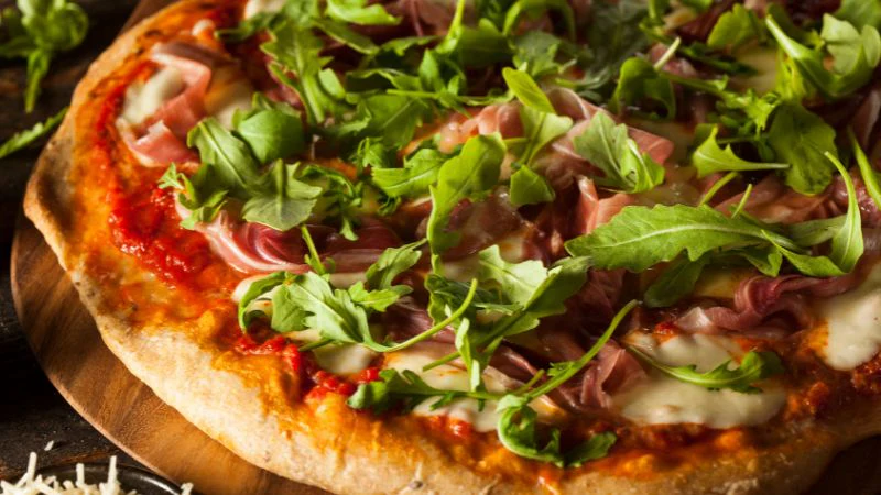 Close-up of a pizza with arugula, prosciutto, and melted cheese on a wooden board.
