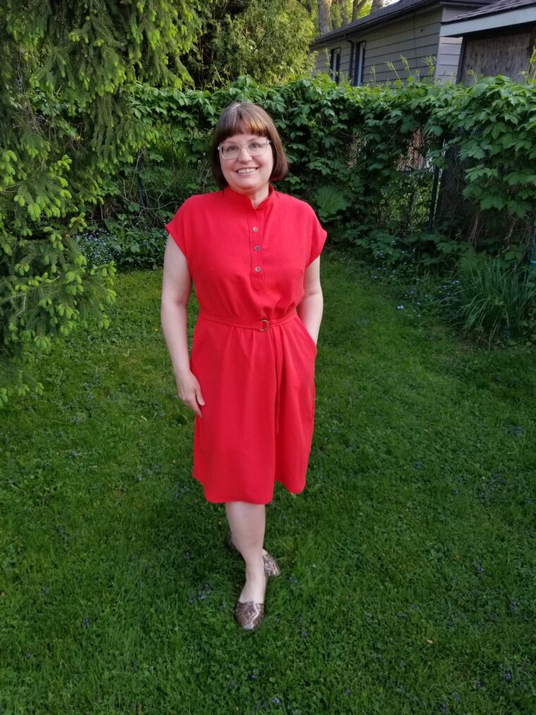 Woman standing in a backyard and smiling, wearing a red short sleeve pullover dress with a button placket at the neckline. She has a belt made in the same fabric.