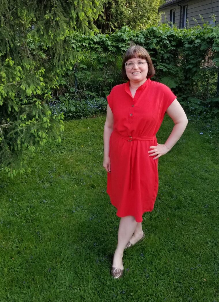 Woman standing in a backyard and smiling, wearing a red short sleeve pullover dress with a button placket at the neckline. She has a belt made in the same fabric.