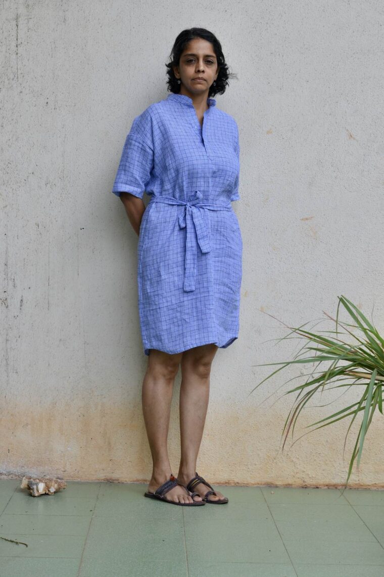A woman wearing the blue linen Santa Rosa Dress. The dress is cinched with a belt. Sheâ€™s standing outside in front of a white wall.