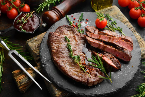 A slate platter with grilled steak slices, herbs, and peppercorns, surrounded by tomatoes, rosemary, a meat fork, and a pepper grinder.