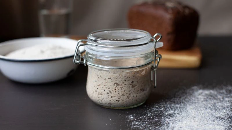 A sourdough starter jar sits on a dark counter with flour and bread in the background.