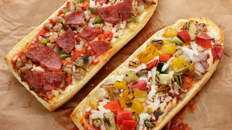 Two rectangular pizza slices on parchment: one with pepperoni, onions, green peppers; the other with assorted veggies.