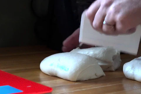 A hand with a ring uses a dough scraper to shape dough on a wooden surface.