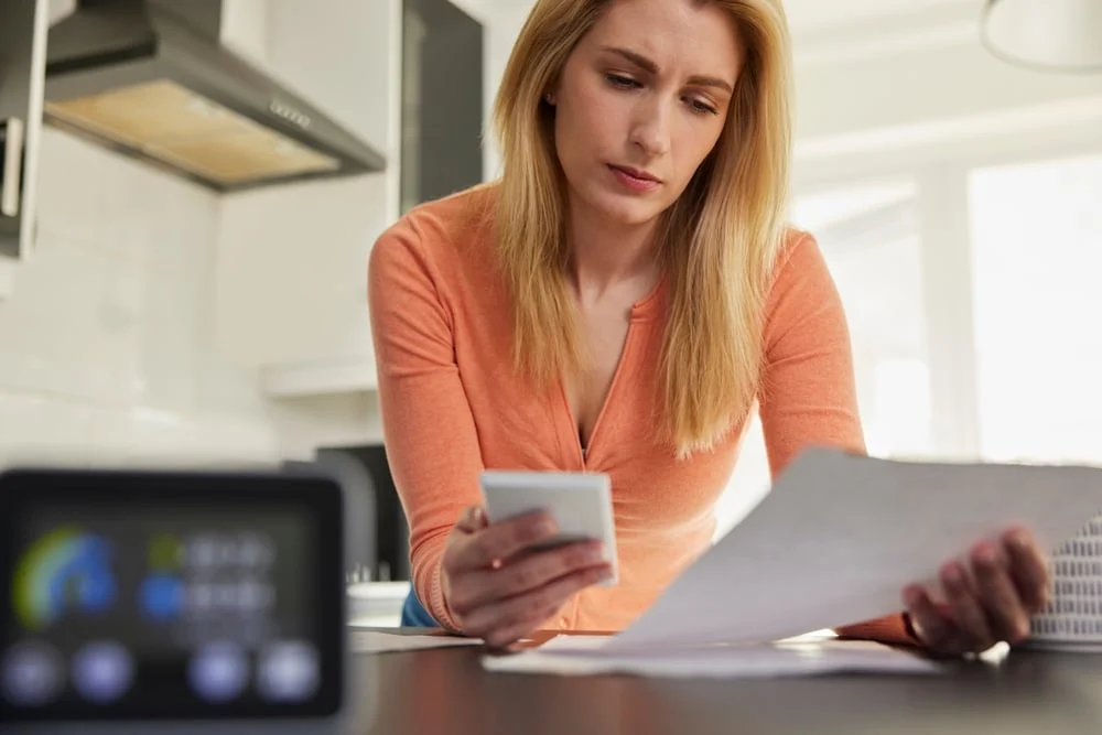 A woman is sitting in her kitchen and is checking her electricity bill, while holding a smartphone.