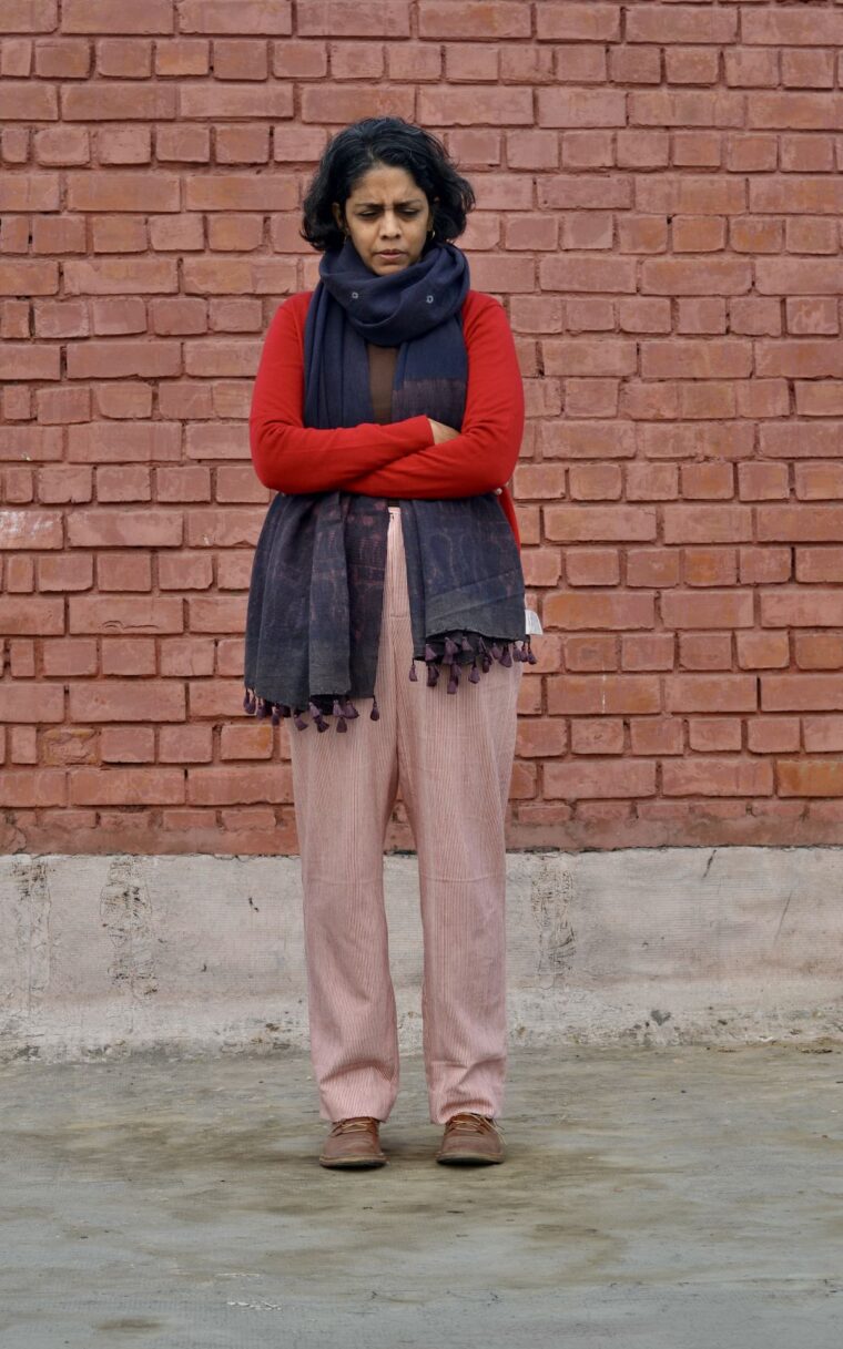 A woman standing in front of a brick wall wearing red and white trousers, a red sweater, and a long scarf. She looks down at the ground in front of her.