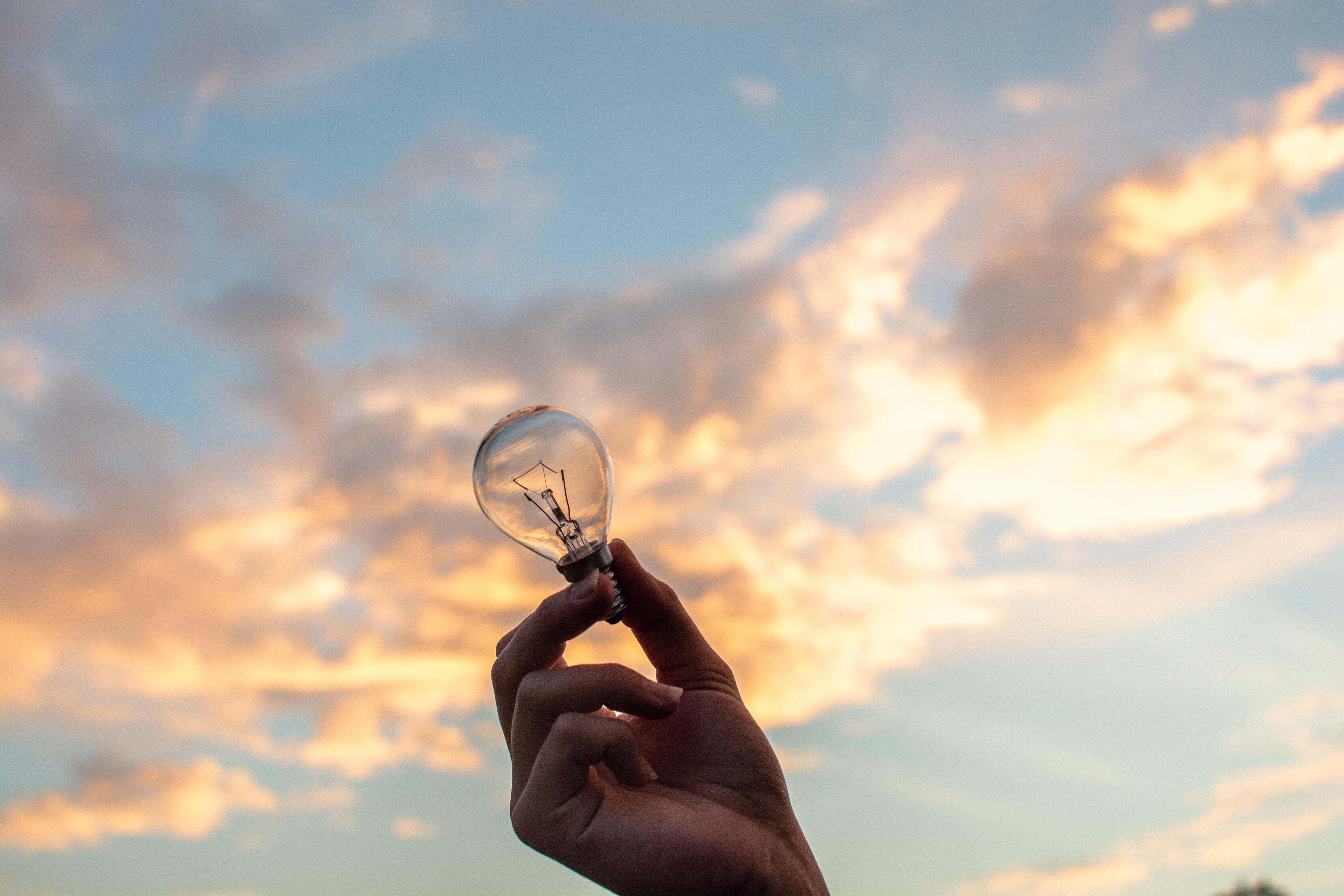 A close-up of a hand holding a light bulb toward the sky during a sunset.