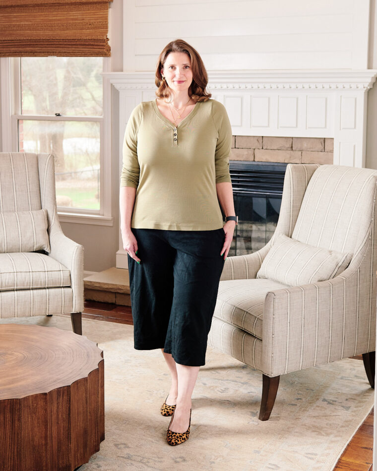 A woman smiling and standing near a fireplace wearing an olive Henley, black cropped pants, and leopard-print pumps.