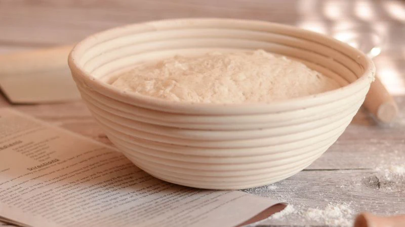 A dough-filled bowl rises on a wooden surface, surrounded by a recipe and kitchen tools.