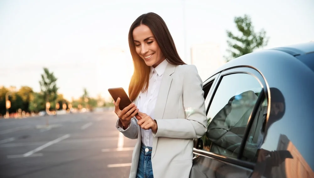 A woman looks at her smart phone as she leans on the side of her EV.
