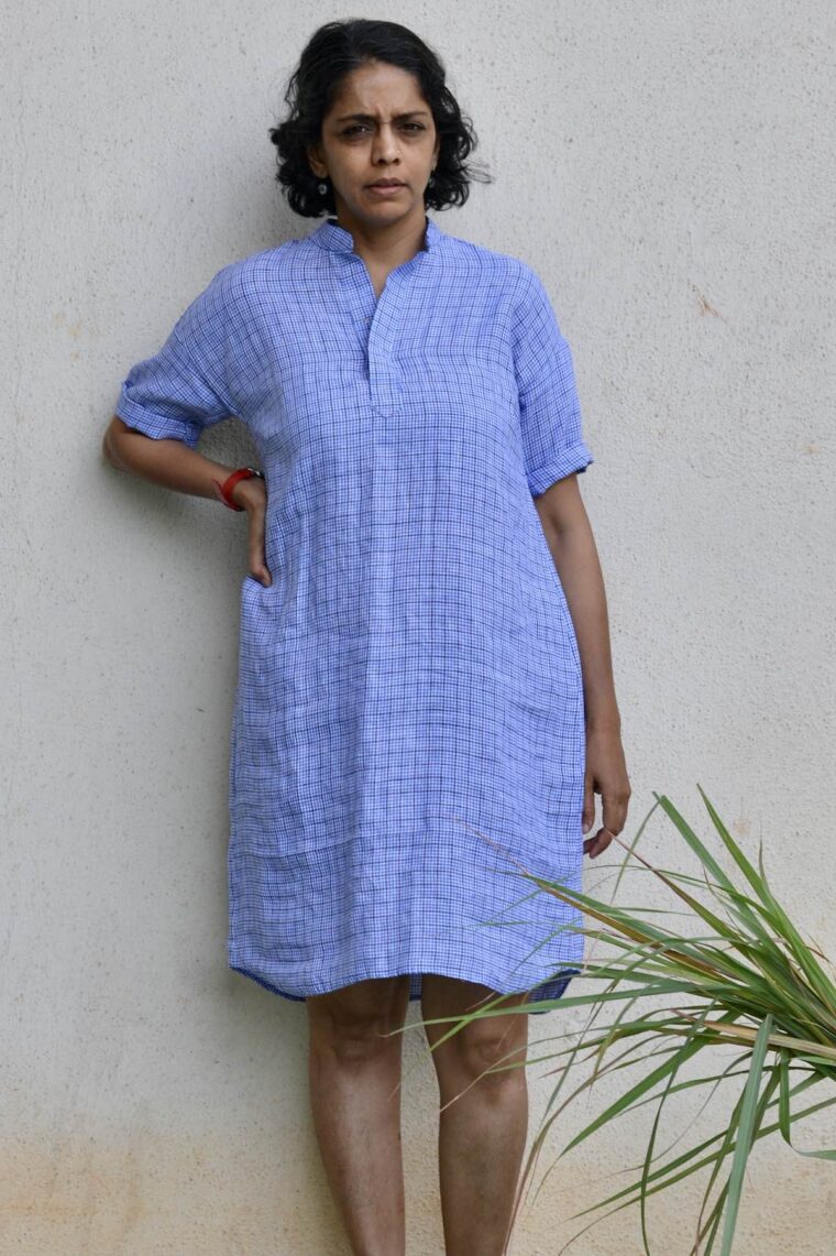 A woman wearing the blue linen Santa Rosa Dress. Sheâ€™s standing outside in front of a white wall.