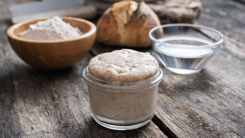 Sourdough starter jar on a table with flour, water, and a loaf of bread in the background.