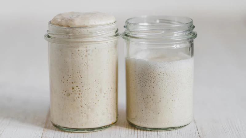 Two glass jars hold dough at different fermentation stages. The left jar's dough is more risen and bubbly than the right. They sit on a white surface.