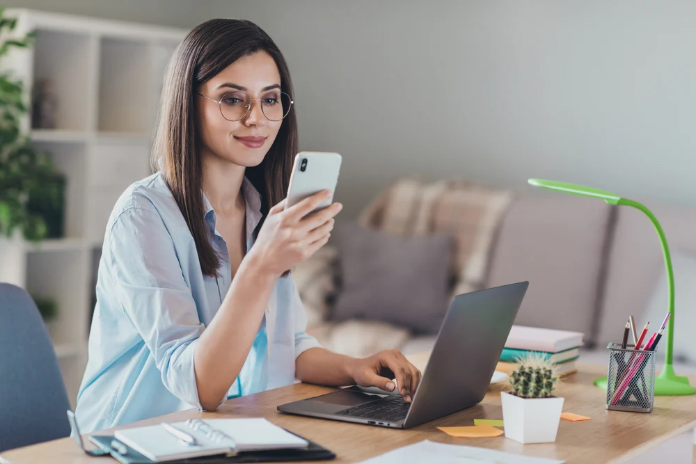 A woman is checking her smartphone while sitting by a desk and working on her laptop.