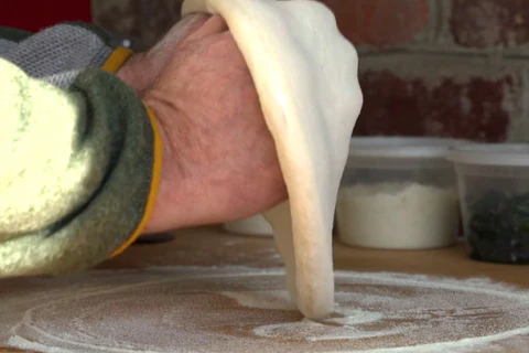 A person stretches pizza dough on a floured surface, ingredients in containers in the background.