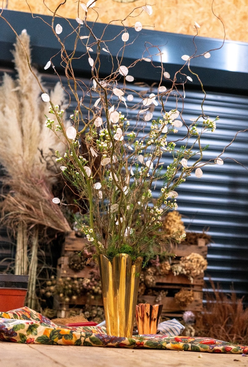 Our Smooth Brass Flora vase with an extravagant arrangement of flowers and dried seeds inside.