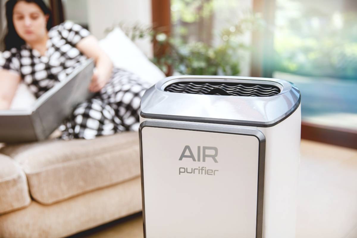 A close-up of a white air purifier in the foreground with a woman reading on a couch in the background, emphasizing the Health Benefits of Air Purifiers for Dogs