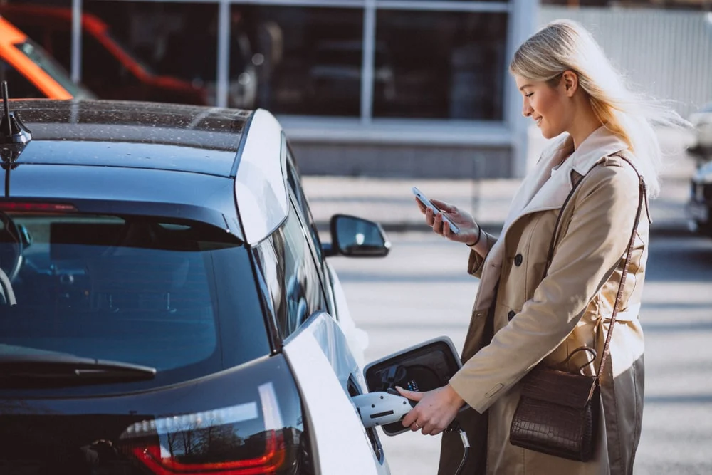 A woman charging her electric car while checking her phone.