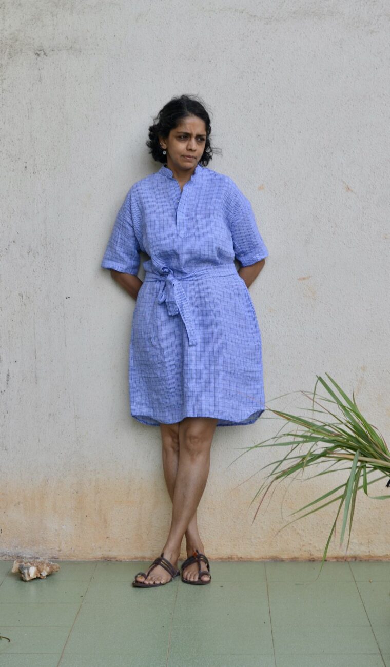 A woman wearing the blue linen Santa Rosa Dress. The dress is cinched with a belt. Sheâ€™s standing outside in front of a white wall.