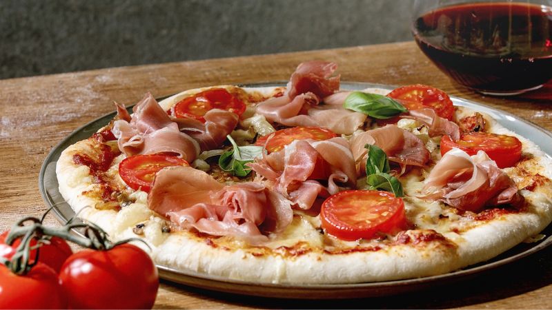 A pizza with tomatoes, prosciutto, basil, and cheese sits on a wooden table beside red wine and fresh tomatoes.