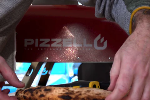 Person handling a wood-fired pizza at a Pizzello red oven.