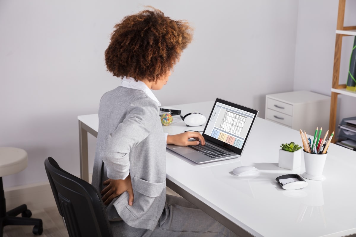 Office worker holding her lower back in pain, demonstrating the importance of using ergonomic seating to enhance comfort during long working hours.