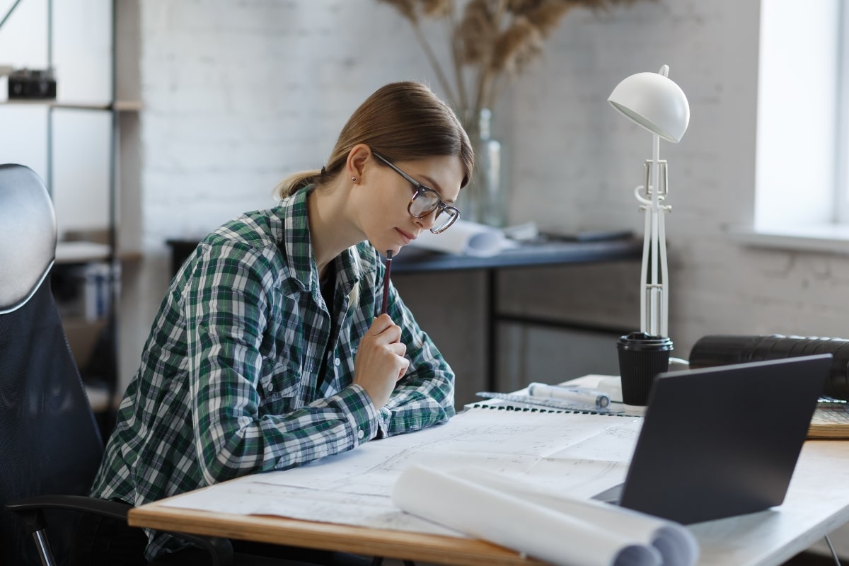 Woman reviewing architectural plans at a desk, demonstrating the ergonomic support and height advantage provided by drafting chairs in design-focused jobs.