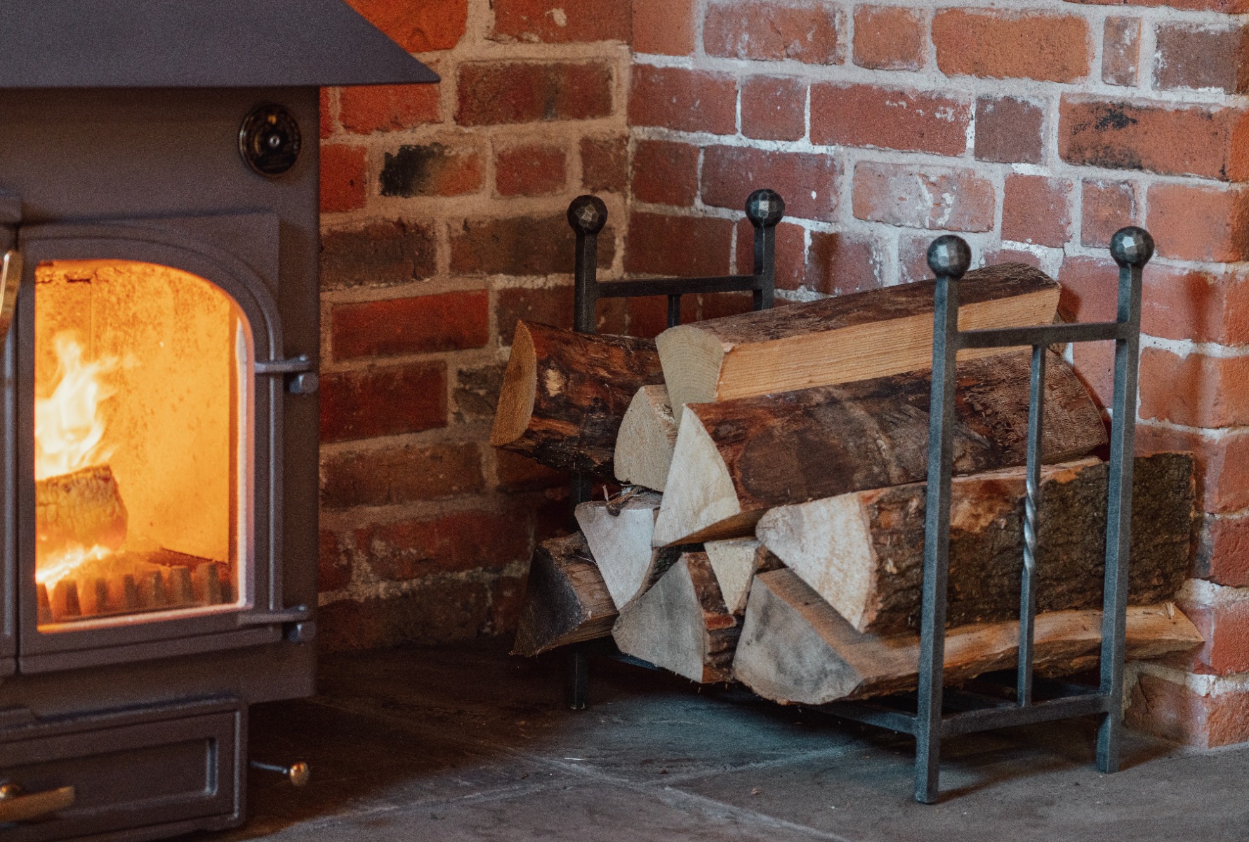 A solid pewter log holder filled with firewood next to a lit log burner.
