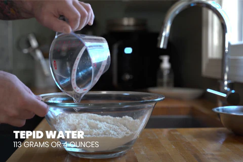 Someone pours 113 grams (4 ounces) of tepid water into a bowl of flour in the kitchen.