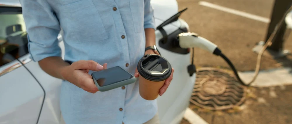 A woman charging her car while checking her phone and holding a take-away coffee cup.