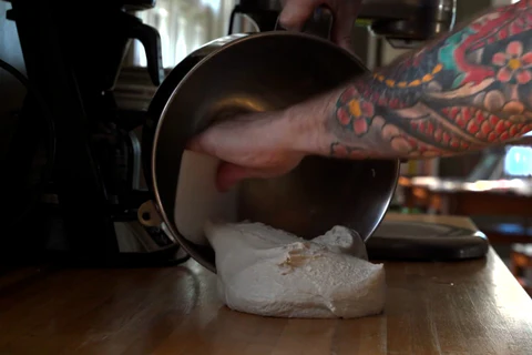 A person scraping dough from a metal mixing bowl onto a wooden surface in the kitchen.