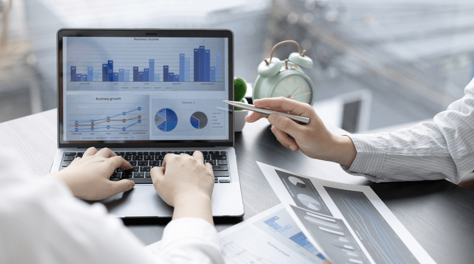 Two hands typing on a laptop while another hand holds a pencil and points at the screen. On the screen, there are several graphs.