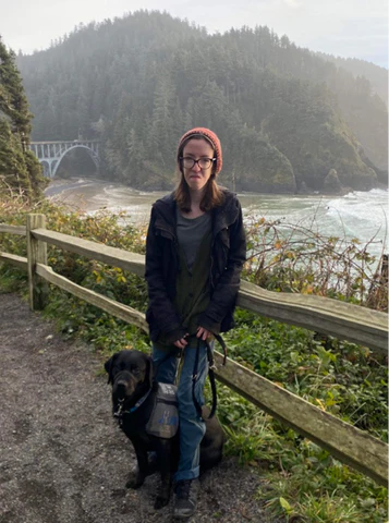 Mary is standing beside her service dog, a black lab, in front of a fence. They are in front of a large body of water and dense trees.