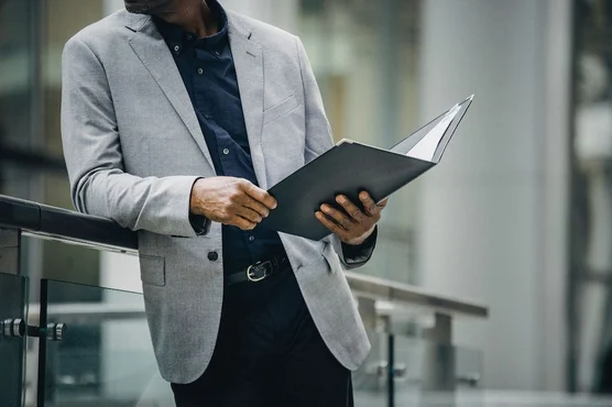A business manager standing on a bridge looking outside while holding a report in his hands.