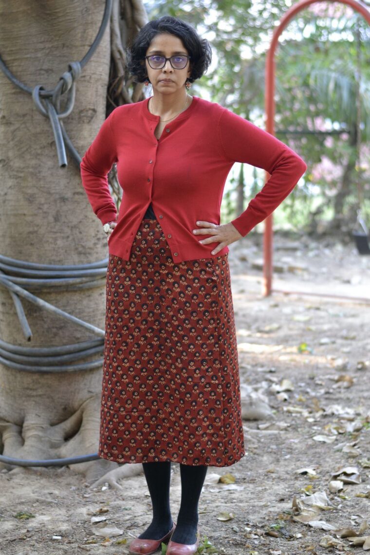 A woman standing wearing a red Garibaldi Skirt, a red button-up cardigan, and red flats